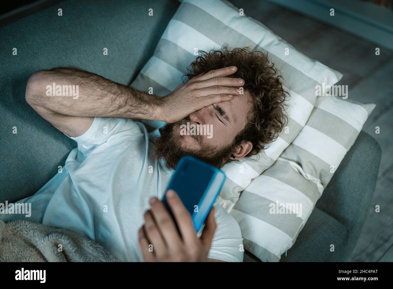 Sleepy Guy désactive l'alarme sur le téléphone.Caucasien Guy se trouve sur le lit et couvre les yeux il a un smartphone dans une main.Appel du matin, alarme du matin, réveil tôt.Gros plan.Arrière-plan de la chambre.Photo de haute qualité Banque D'Images