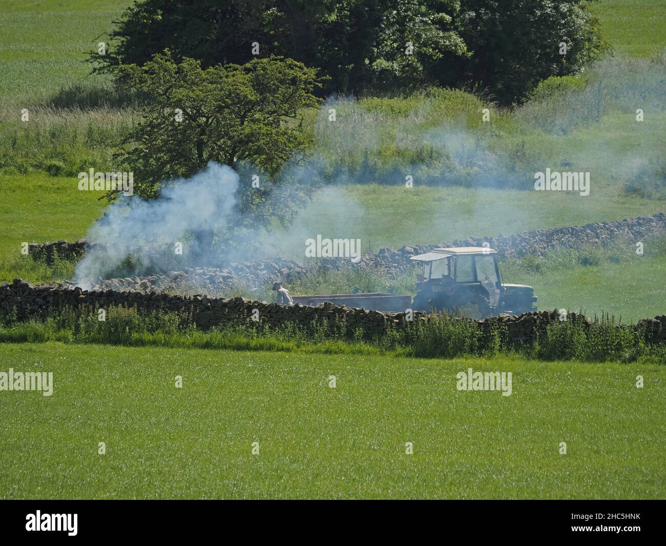 Paysage rural d'été avec des fumées de feu de camp en hausse dans le coin du champ avec agriculteur, tracteur et remorque en présence - Cumbria, Angleterre, Royaume-Uni Banque D'Images