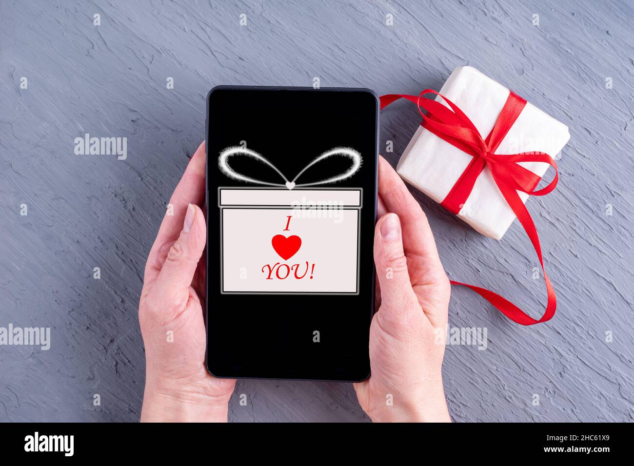 Les femmes tiennent un téléphone avec une icône cadeau et l'inscription Je VOUS AIME et un cadeau sur fond gris.Vente en ligne pour la Saint-Valentin.En ligne Banque D'Images