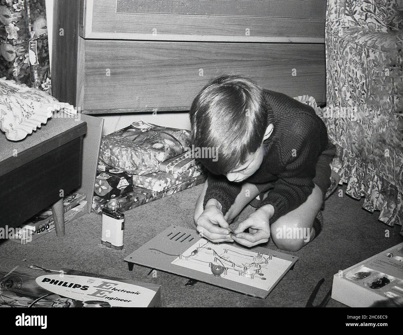 1960s, historique, à l'intérieur d'une pièce, un jeune garçon s'agenouillant sur le sol en jouant avec un cadeau de noël, une carte de circuit imprimé d'un kit d'ingénieur électronique Philips, Angleterre, Royaume-Uni.Les kits de « jeunes ingénieurs » en boîte étaient une activité éducative et ludique populaire que les jeunes garçons devaient entreprendre à cette époque. Banque D'Images