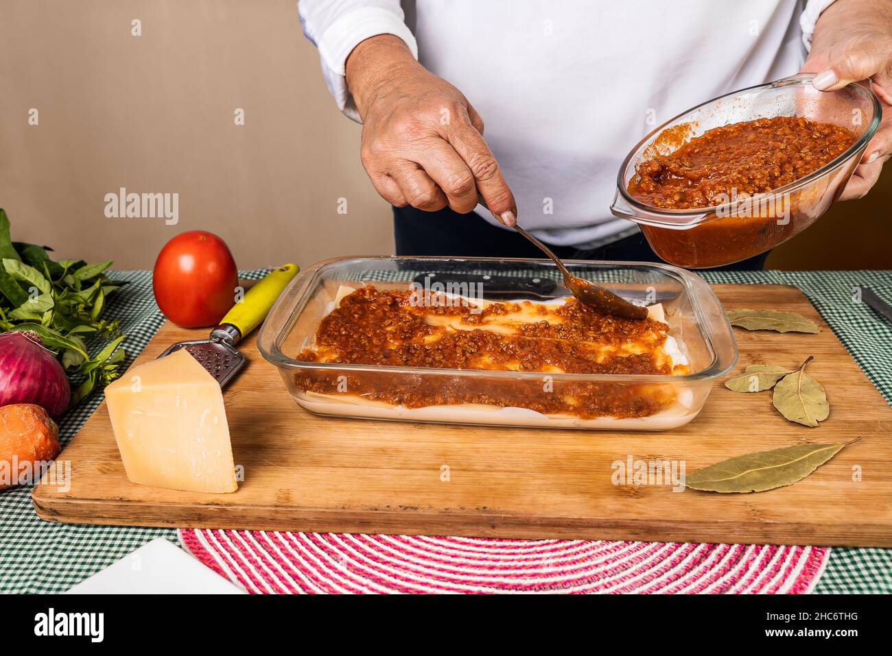 Une femme méconnaissable qui prépare une recette de lasagnes en plaçant la sauce bolognaise sur la préparation avec les différents ingrédients autour du contai Banque D'Images