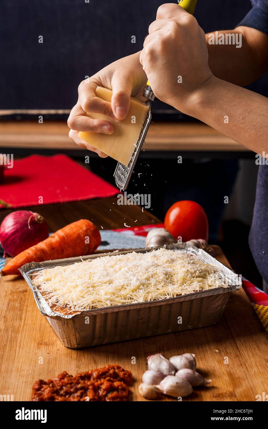 Image verticale d'une personne méconnaissable qui râpe du fromage frais sur des lasagnes avec une râpe avec différents types d'ingrédients autour de la nourriture. Banque D'Images