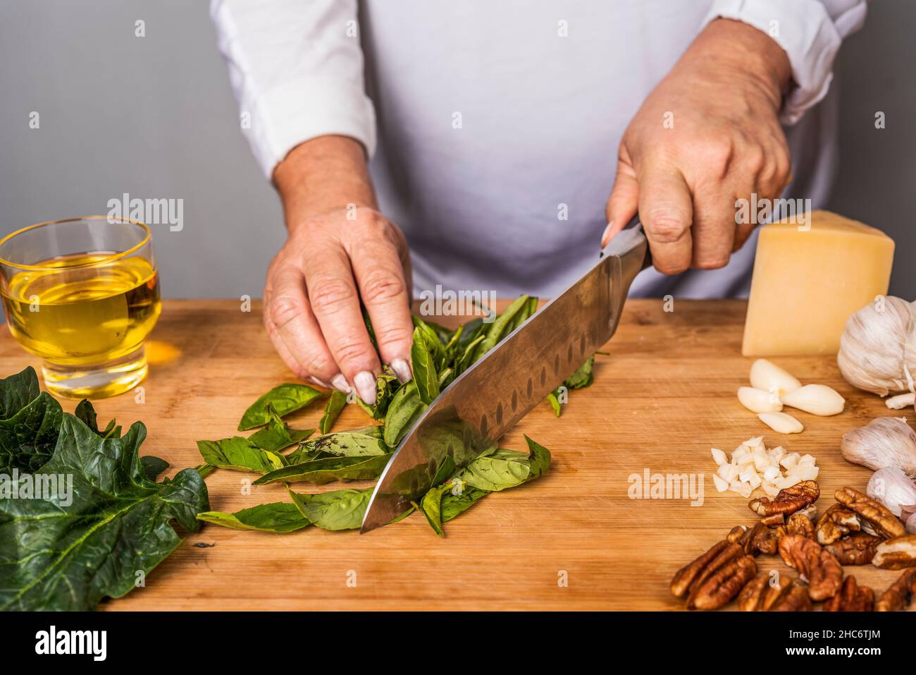 Vue rognée d'une femme adulte méconnue qui hache du basilic avec un couteau de cuisine sur une table en bois pour la préparation d'une sauce au pesto. Banque D'Images