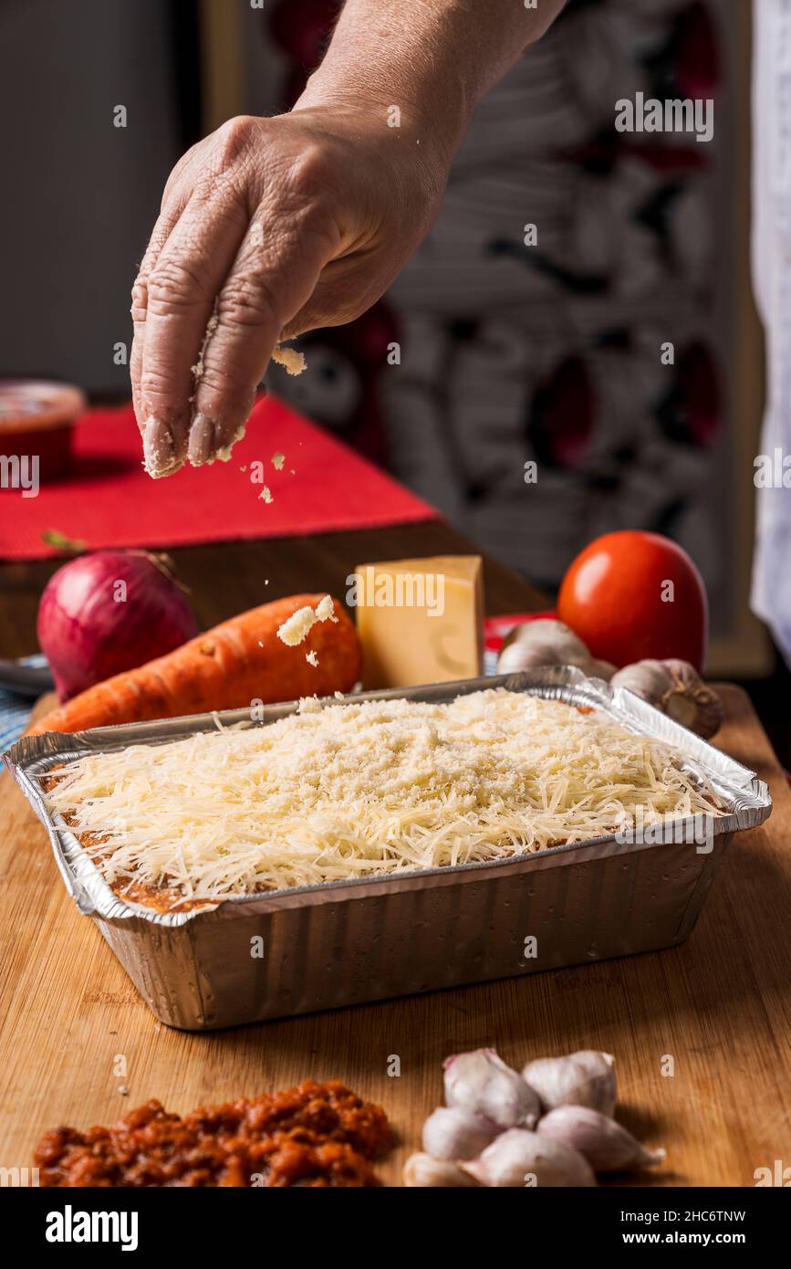 Image verticale d'une main d'une personne méconnaissable versant beaucoup de fromage râpé sur des lasagnes avec différents types d'ingrédients autour d'elle. Banque D'Images