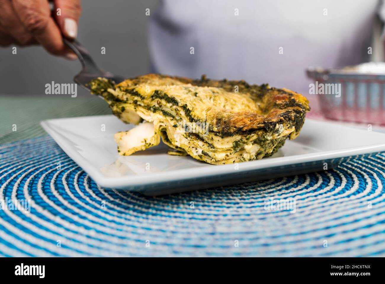 Vue rapprochée d'une main d'une femme adulte servant une portion de lasagne au pesto fraîchement préparée sur une assiette. Banque D'Images