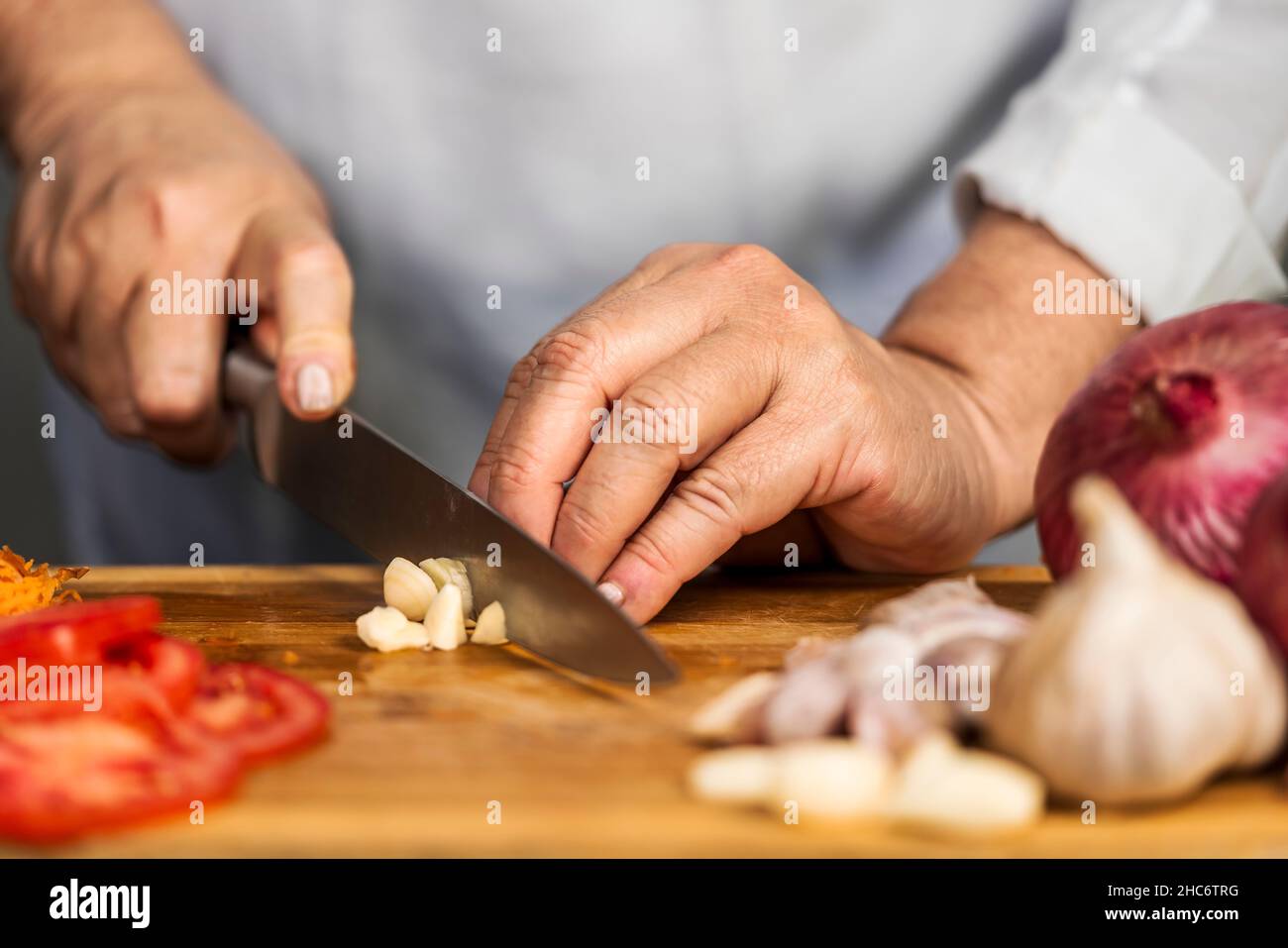 Vue rapprochée des mains d'une femme adulte hacher un ail avec un couteau de cuisine sur une planche à découper pour la préparation de lasagnes. Banque D'Images
