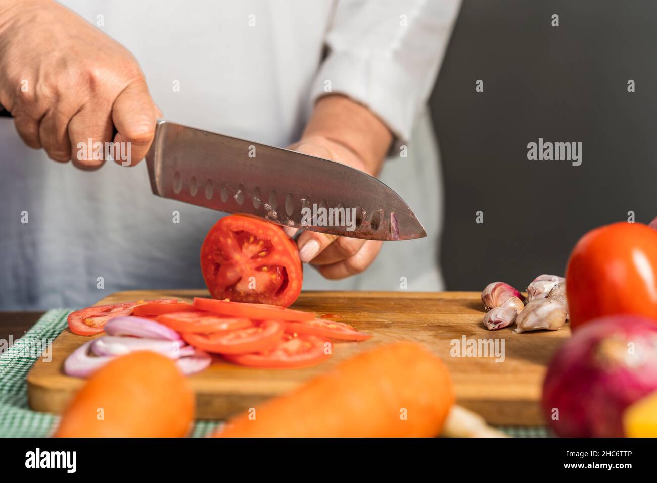 Vue rapprochée des mains d'une femme adulte coupant des tomates en tranches à l'aide d'un couteau de cuisine sur une planche à découper en bois pour la préparation de lasagnes. Banque D'Images