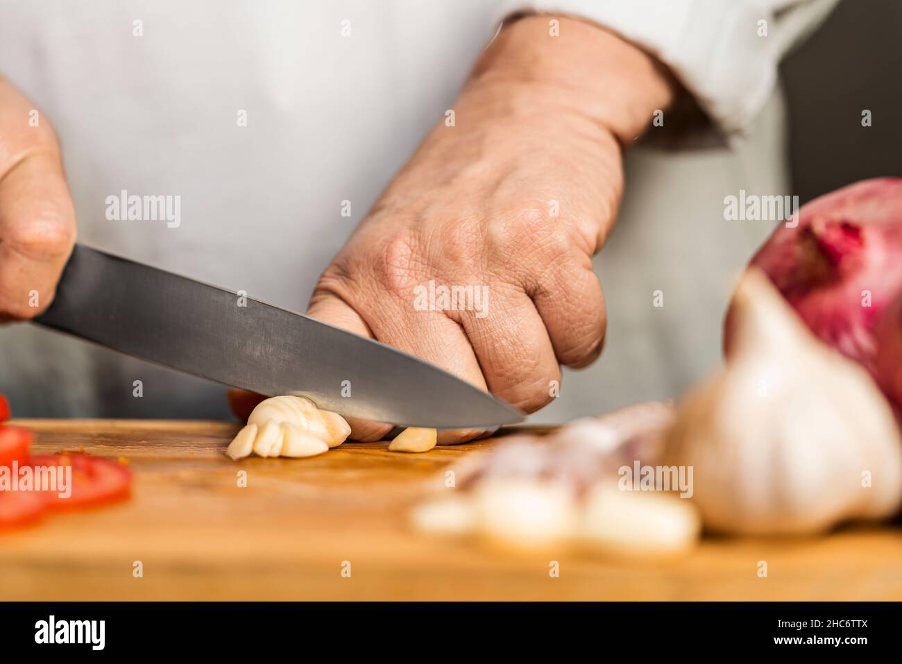 Vue rapprochée des mains d'une femme adulte coupant un ail avec un couteau de cuisine sur une planche à découper pour la préparation de lasagnes. Banque D'Images