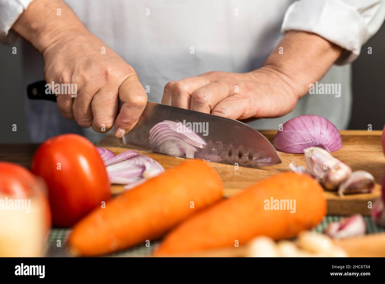 Vue rapprochée des mains d'une femme adulte coupant un oignon avec un couteau de cuisine sur une planche en bois pour la préparation de lasagnes. Banque D'Images