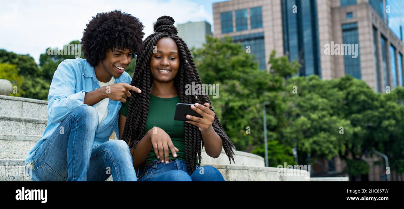 Influenceur afro-américain, homme et femme, publiant un message avec un téléphone mobile en plein air dans la ville en été Banque D'Images