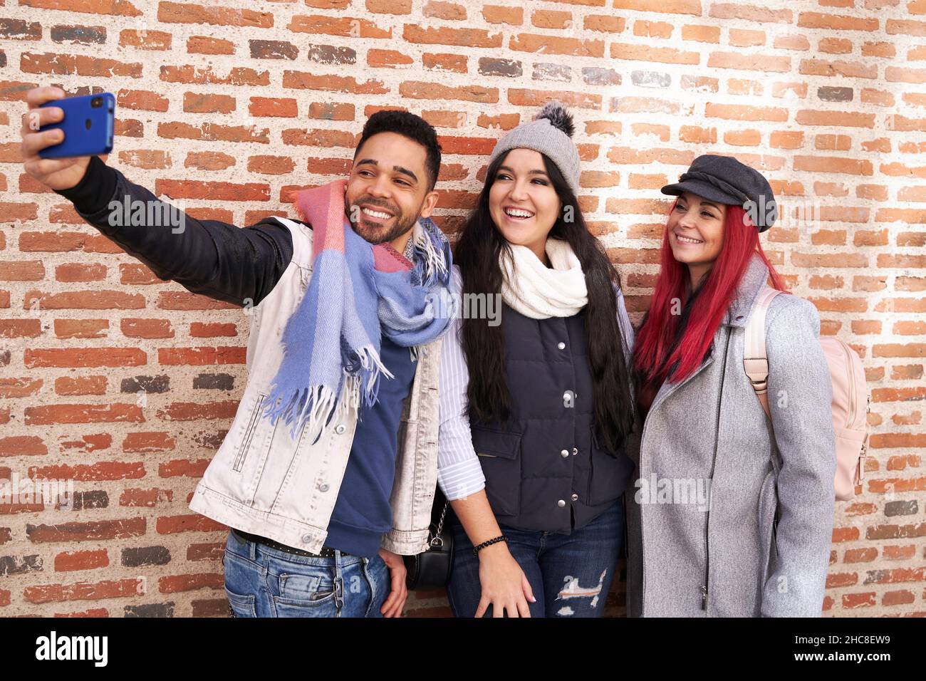 Amis multiraciaux positifs, hommes et femmes, dans des tenues stylées, souriant de façon éclatante tout en prenant un autoportrait sur un téléphone portable près du revêtement de briques Banque D'Images