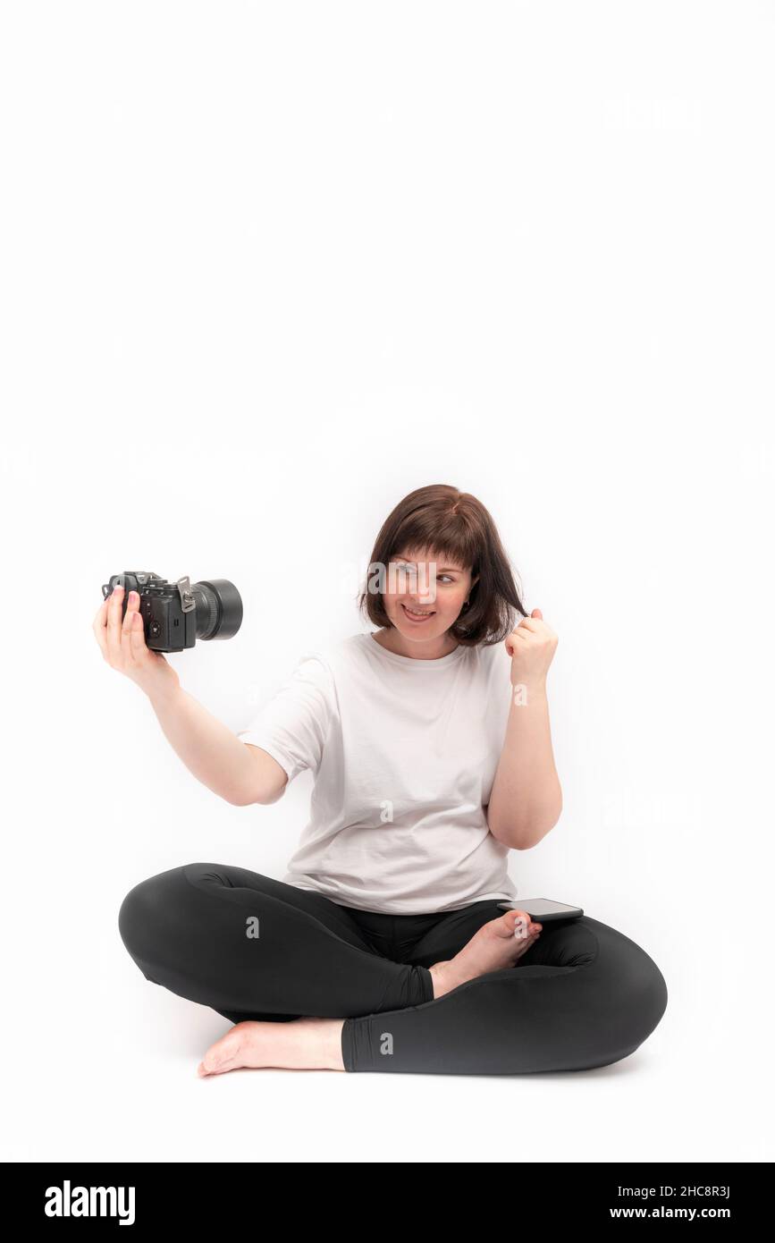 Jeune femme gaie pratique le yoga et prend des photos d'elle-même avec l'appareil photo.Portrait d'une fille dans la moitié de lotus poser sur fond blanc Banque D'Images