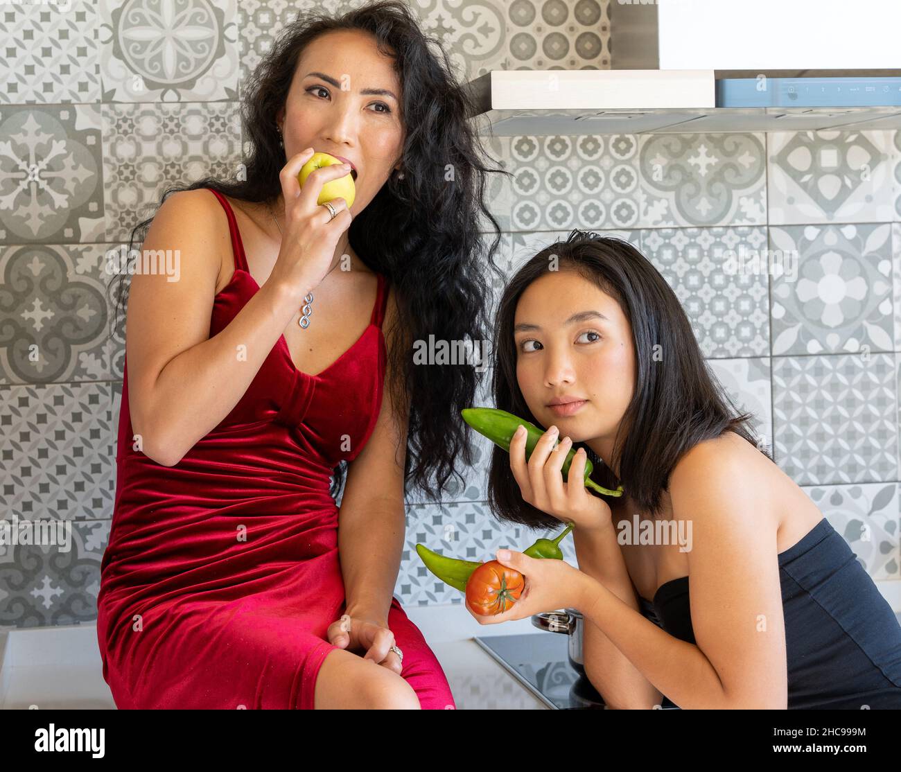 Deux filles asiatiques bien vêtues posant dans la cuisine avec des légumes dans leurs mains, fond carrelé Banque D'Images