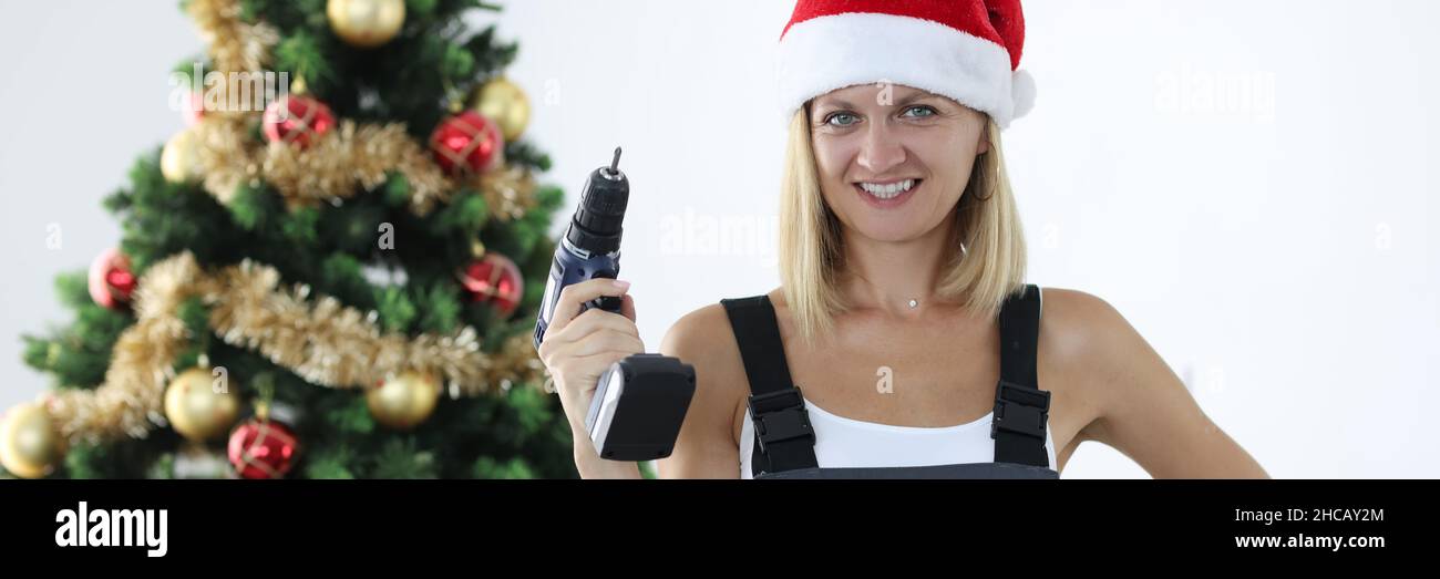 Femme souriante réparateur en uniforme et chapeau du père noël tenant le foret dans les mains près de l'arbre de noël Banque D'Images
