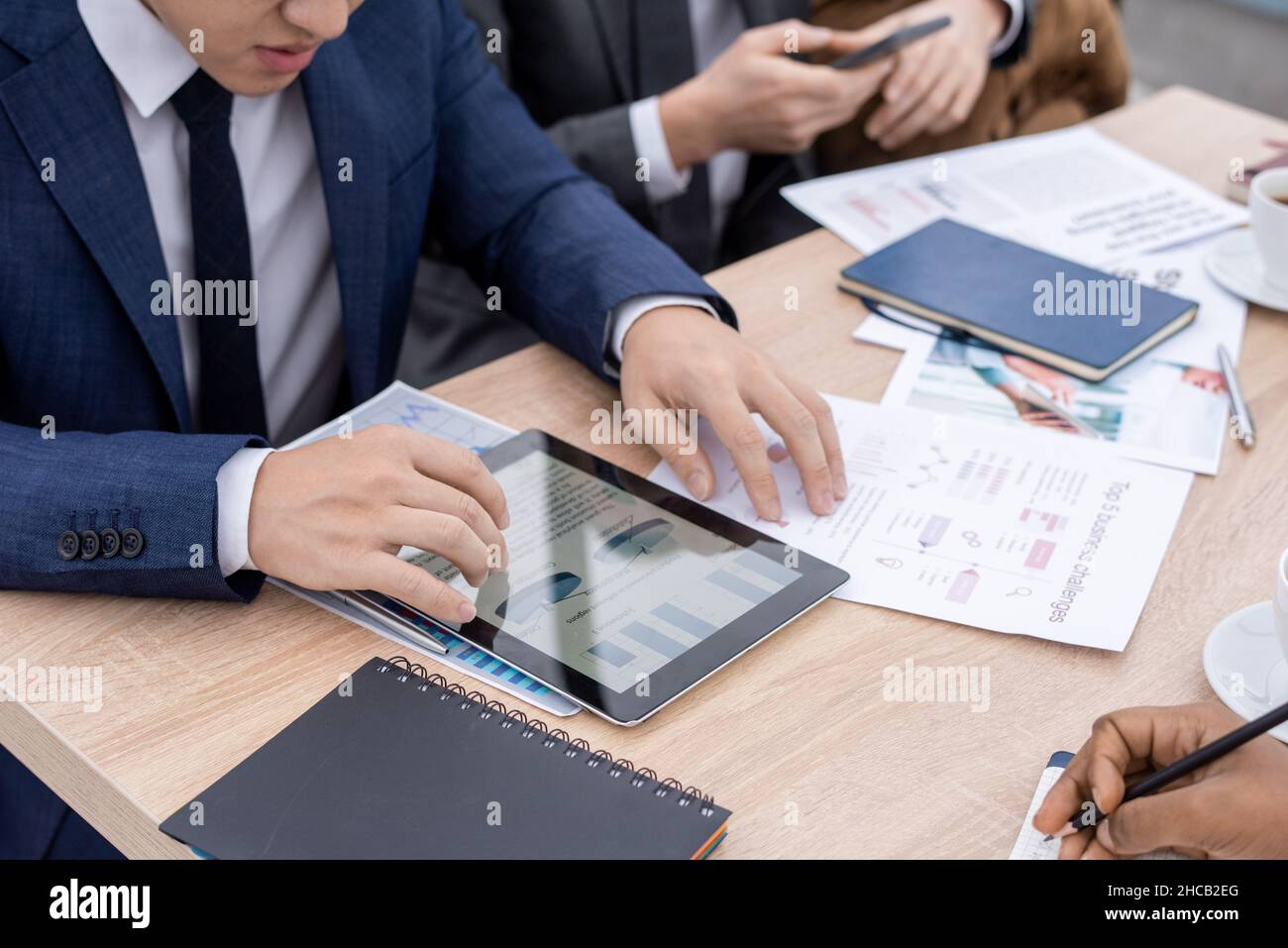 Économiste contemporain en costume regardant à travers les graphiques et les diagrammes en tablette tout en étant assis à table entre collègues Banque D'Images