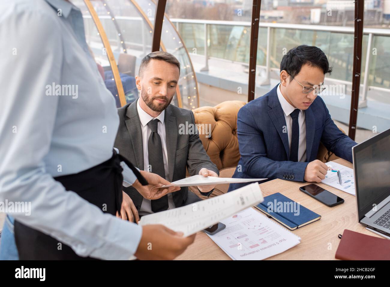 Jeune homme d'affaires sérieux prenant le menu tenu par une serveuse tout en étant assis à table à côté de son partenaire d'affaires chinois Banque D'Images