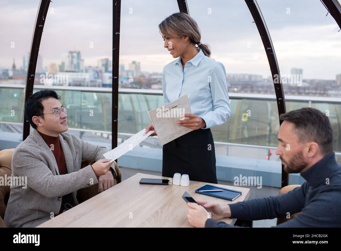 Bonne serveuse jeune passant le menu à l'un des hommes d'affaires interculturels assis à table dans un café ou un restaurant Banque D'Images
