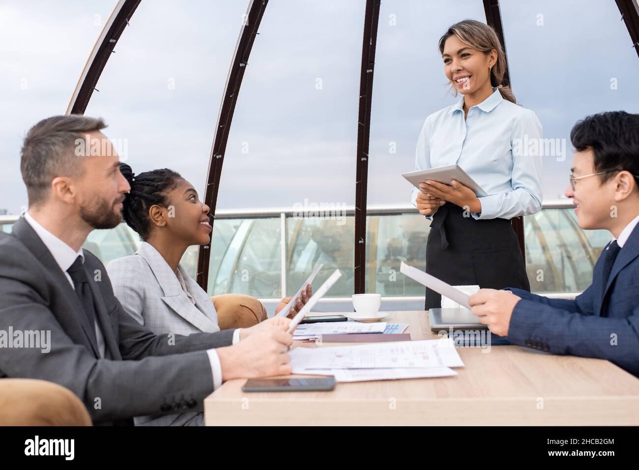 Bonne serveuse jeune avec tablette prenant des commandes de partenaires d'affaires interculturels avec des menus tout en se tenant devant leur table Banque D'Images