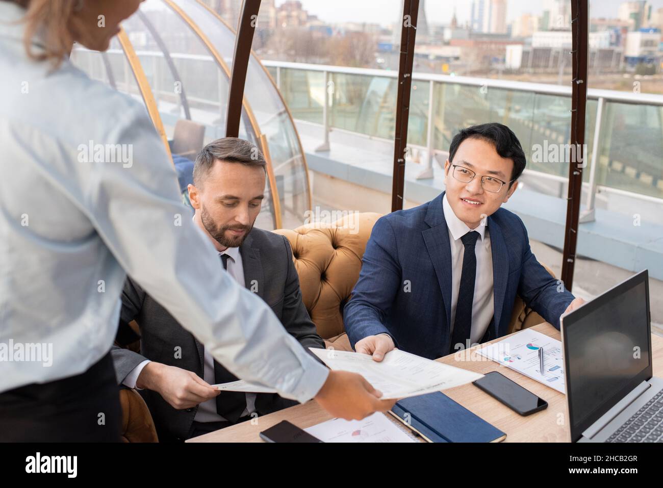 Un homme d'affaires asiatique heureux prenant le menu des mains d'une serveuse et la regardant alors qu'elle était assise à table à côté d'une collègue Banque D'Images