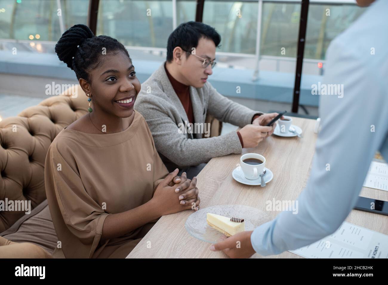Bonne jeune femme africaine en tenue décontractée élégante regardant la plaque de mise de serveuse avec cheesecake sur table dans le café Banque D'Images
