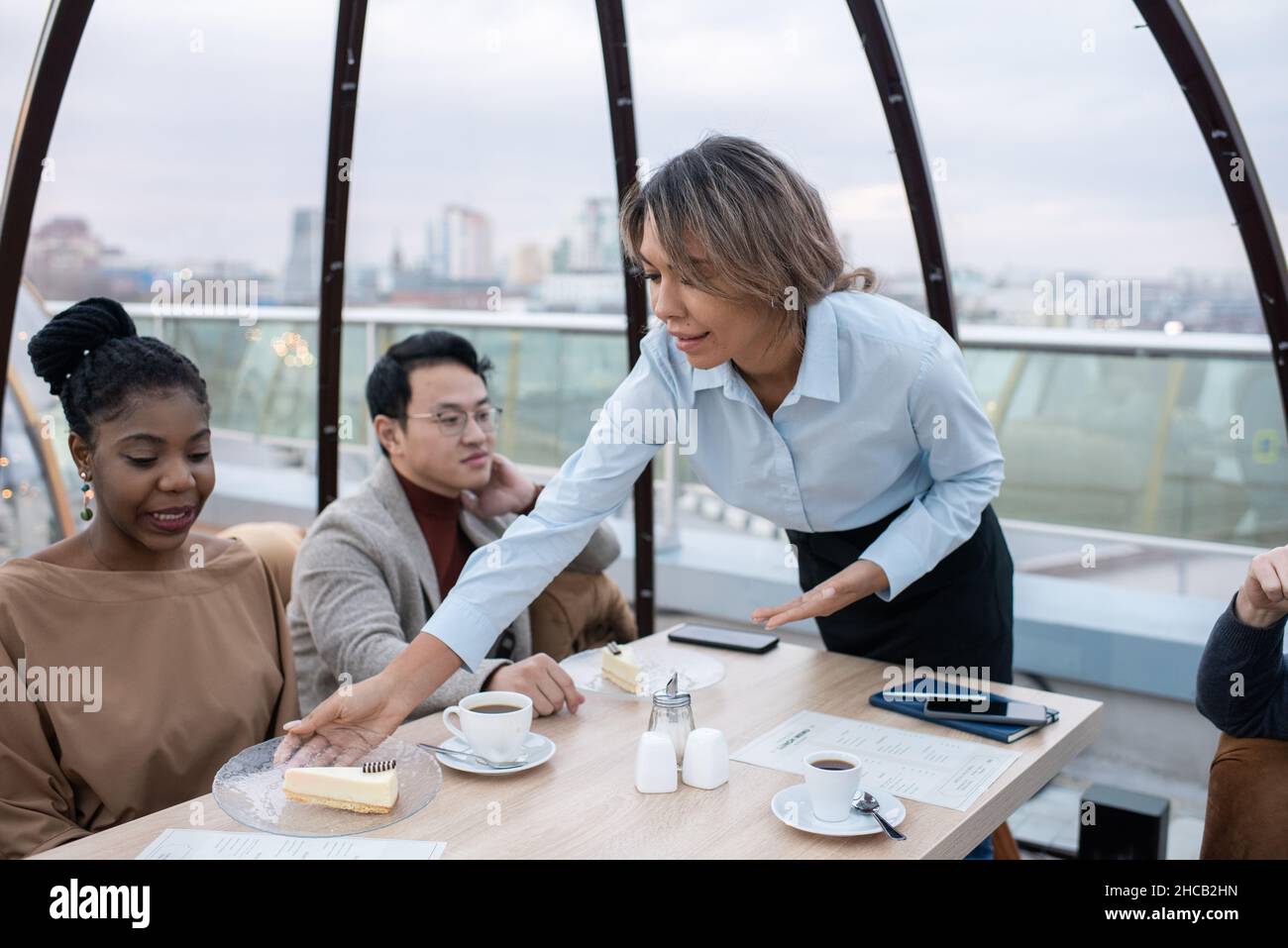 Jeune serveuse se pliant sur une table et une assiette de mise avec un appétissant cheesecake devant une femme africaine Banque D'Images