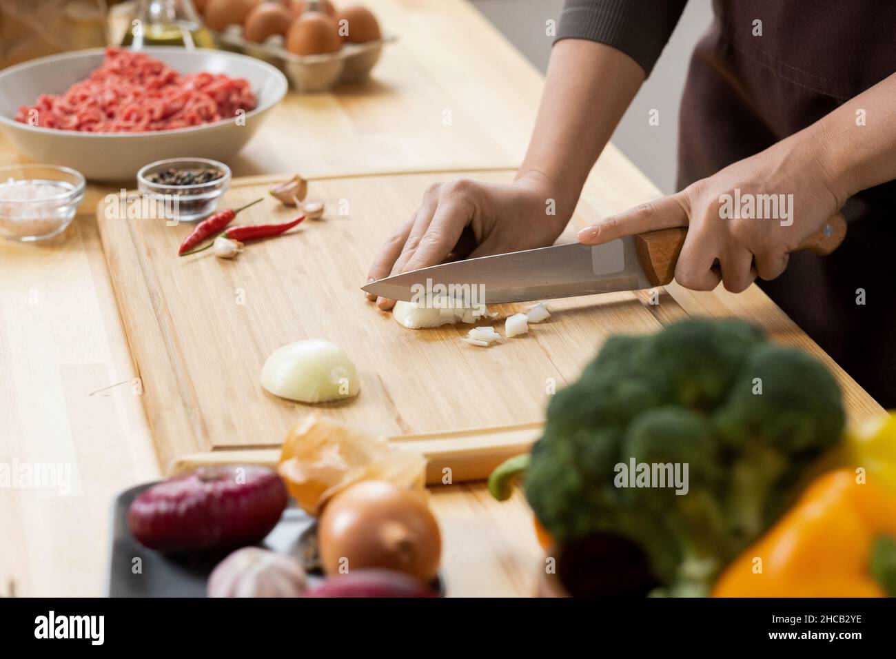 Les mains de la jeune femme coupant l'oignon brut avec un couteau de cuisine aiguisé sur une planche de bois tout en cuisinant de la nourriture Banque D'Images
