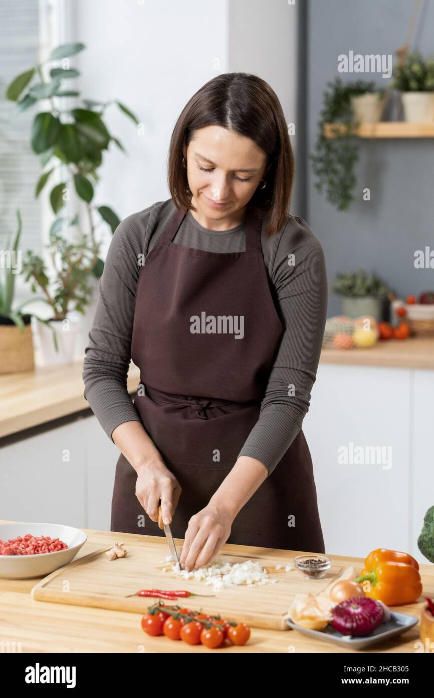 Jeune jolie femme coupant l'oignon sur une planche en bois tout en se tenant près de la table de cuisine et de la cuisine pour le dîner Banque D'Images