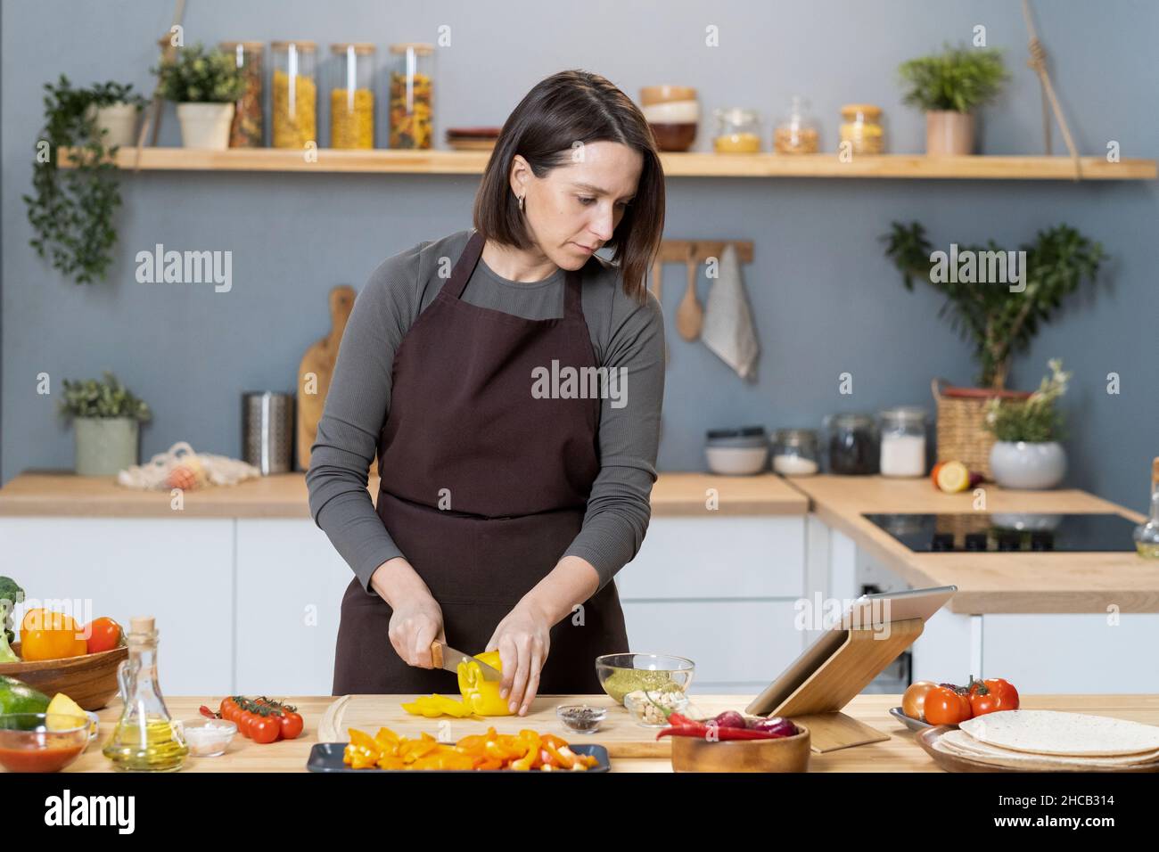 Jeune femme en tablier regardant la recette vidéo tout en coupant le capsicum par la table de cuisine dans l'environnement de la maison Banque D'Images