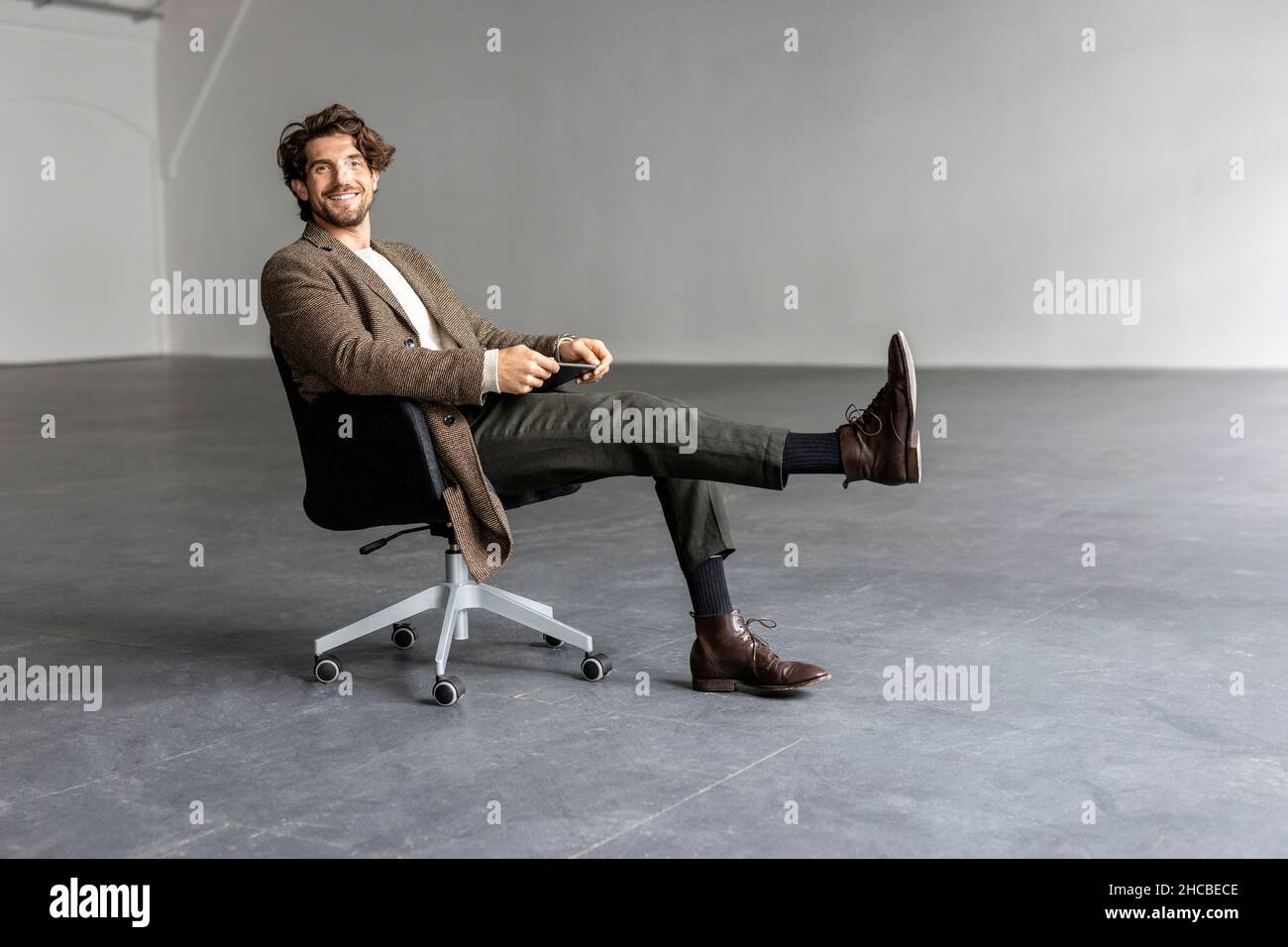 Homme d'affaires souriant avec le pied relevé assis sur une chaise dans une salle industrielle vide Banque D'Images