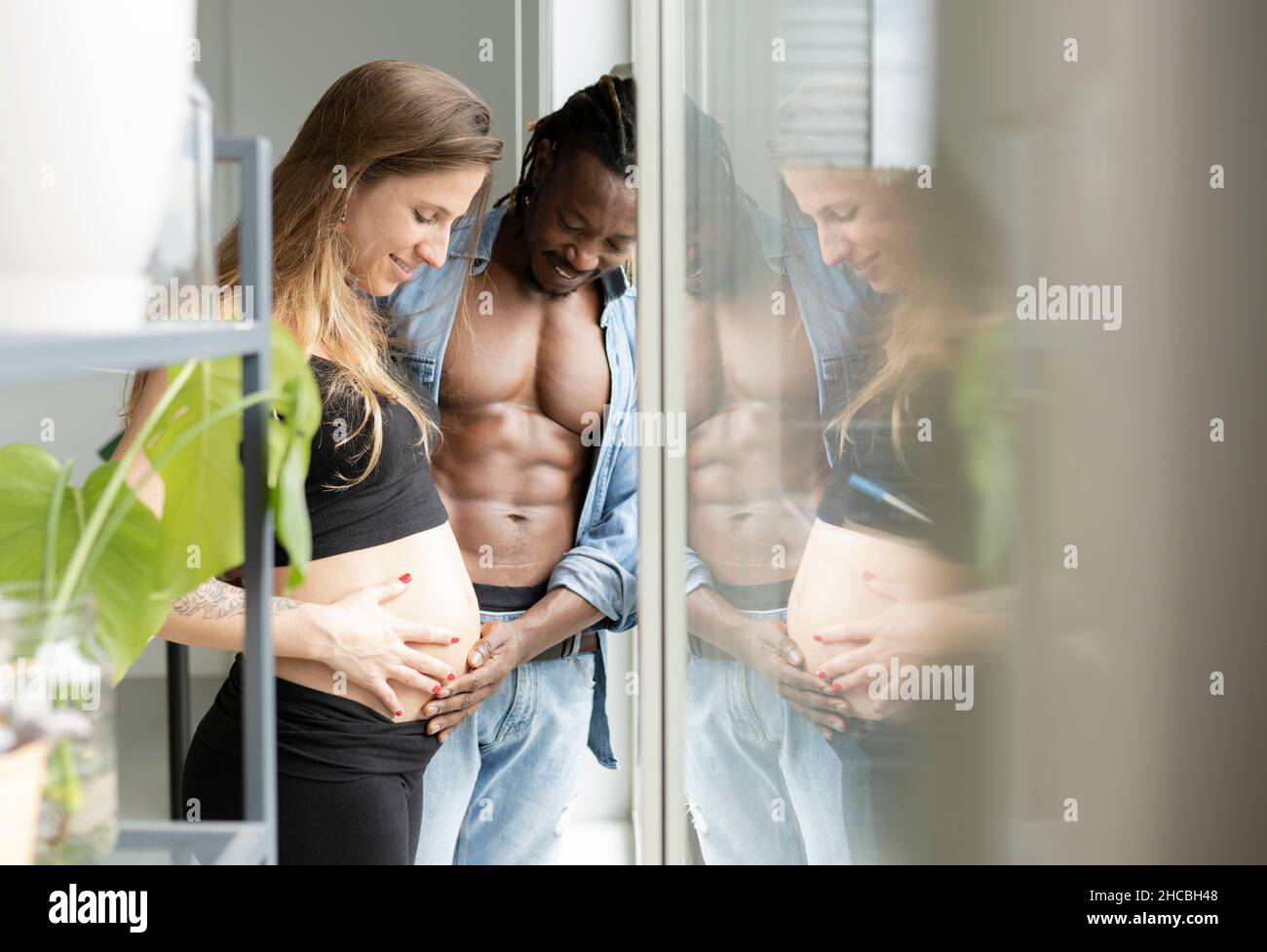 Couple souriant avec les mains sur le ventre regardant par la fenêtre à la maison Banque D'Images