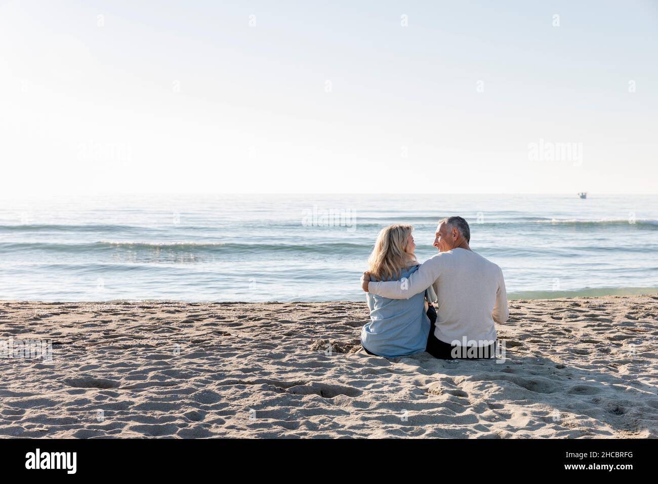 Homme avec le bras autour de la femme assis ensemble sur le sable à la plage Banque D'Images
