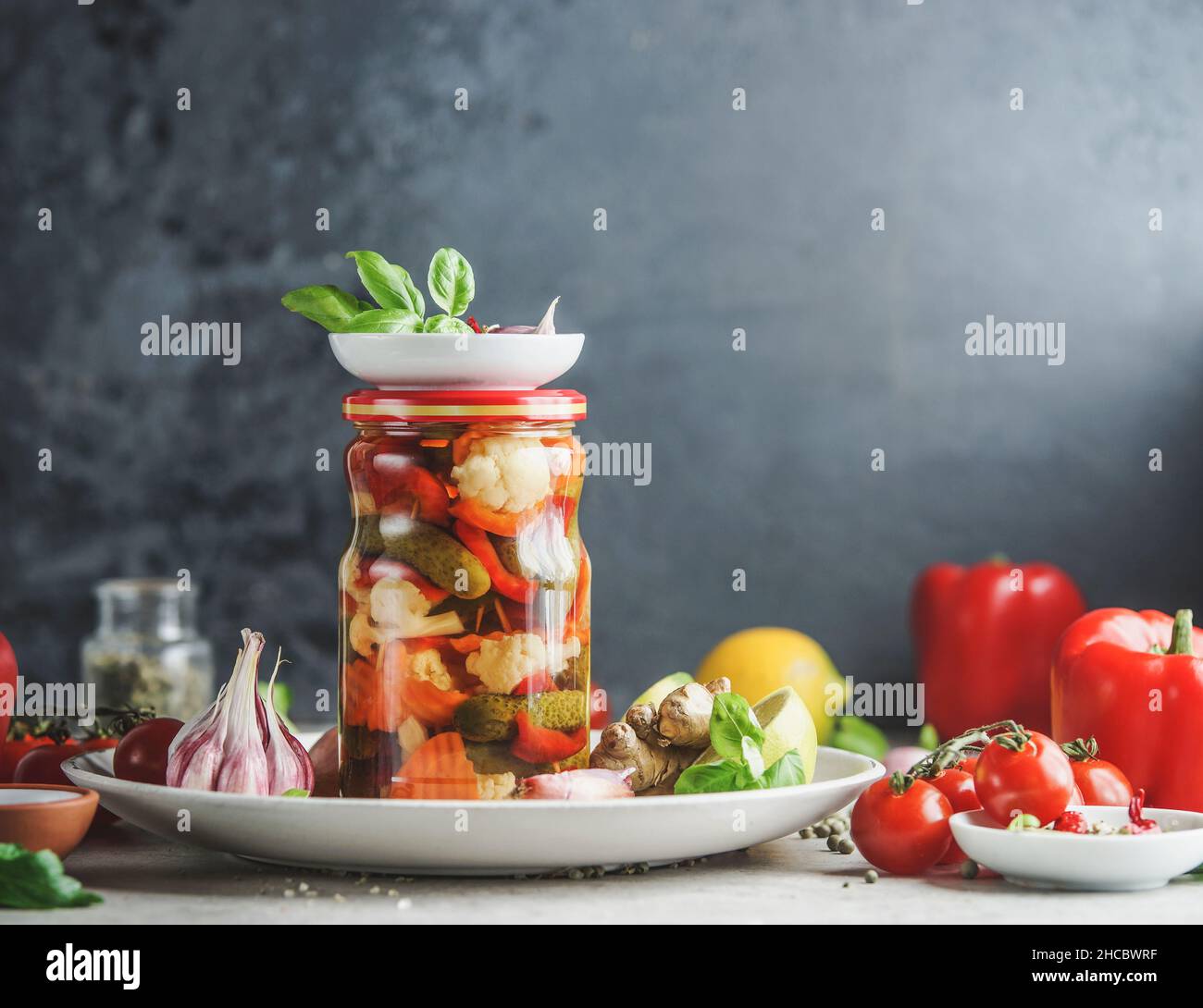 Légumes conservés maison dans un pot en verre sur une plaque blanche à la table avec ustensiles et ingrédients de cuisine, herbes et légumes au fond de mur noir Banque D'Images