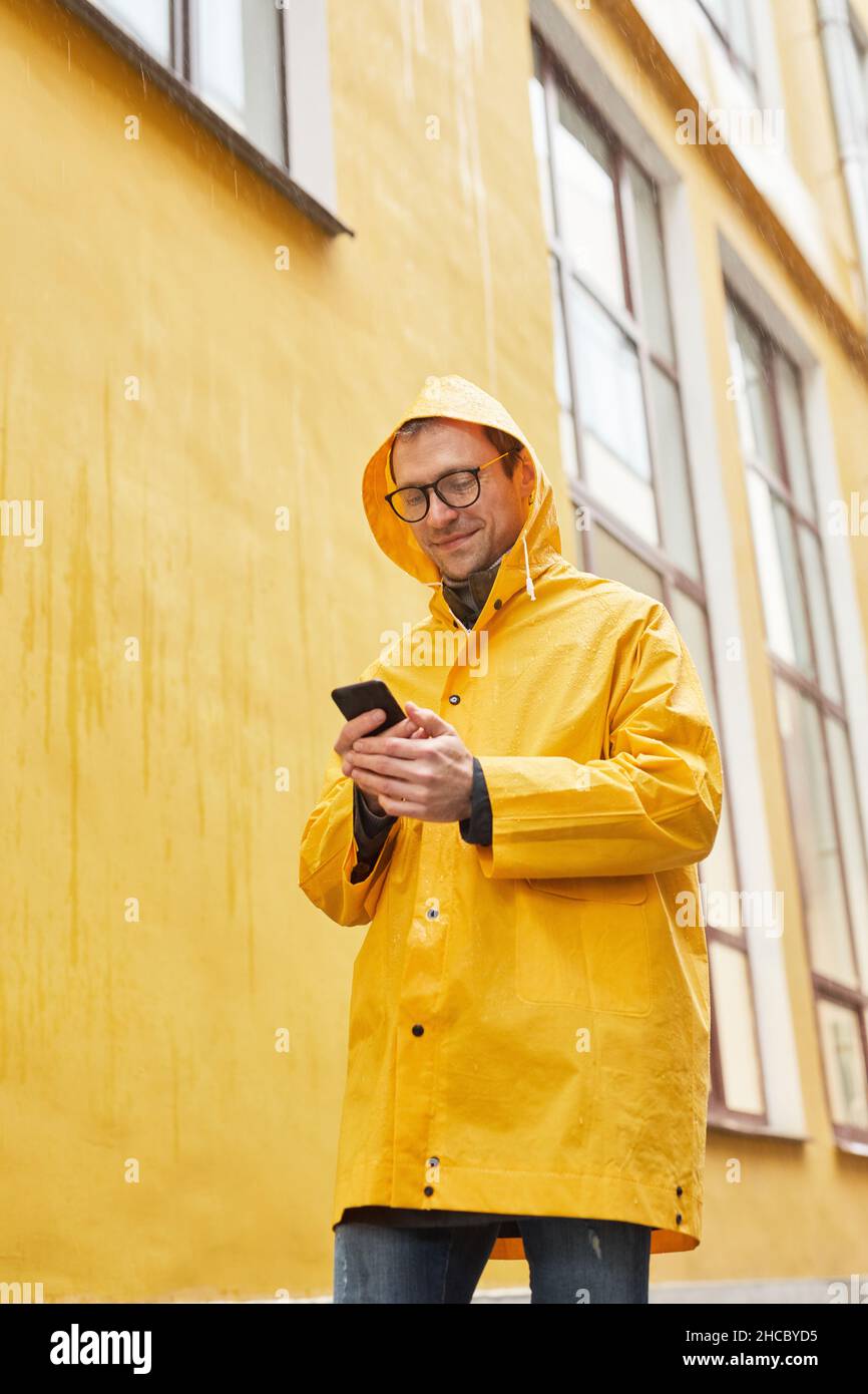Prise de vue verticale à faible angle d'un homme caucasien mature portant un imperméable jaune marchant sous la pluie à l'aide d'un smartphone Banque D'Images
