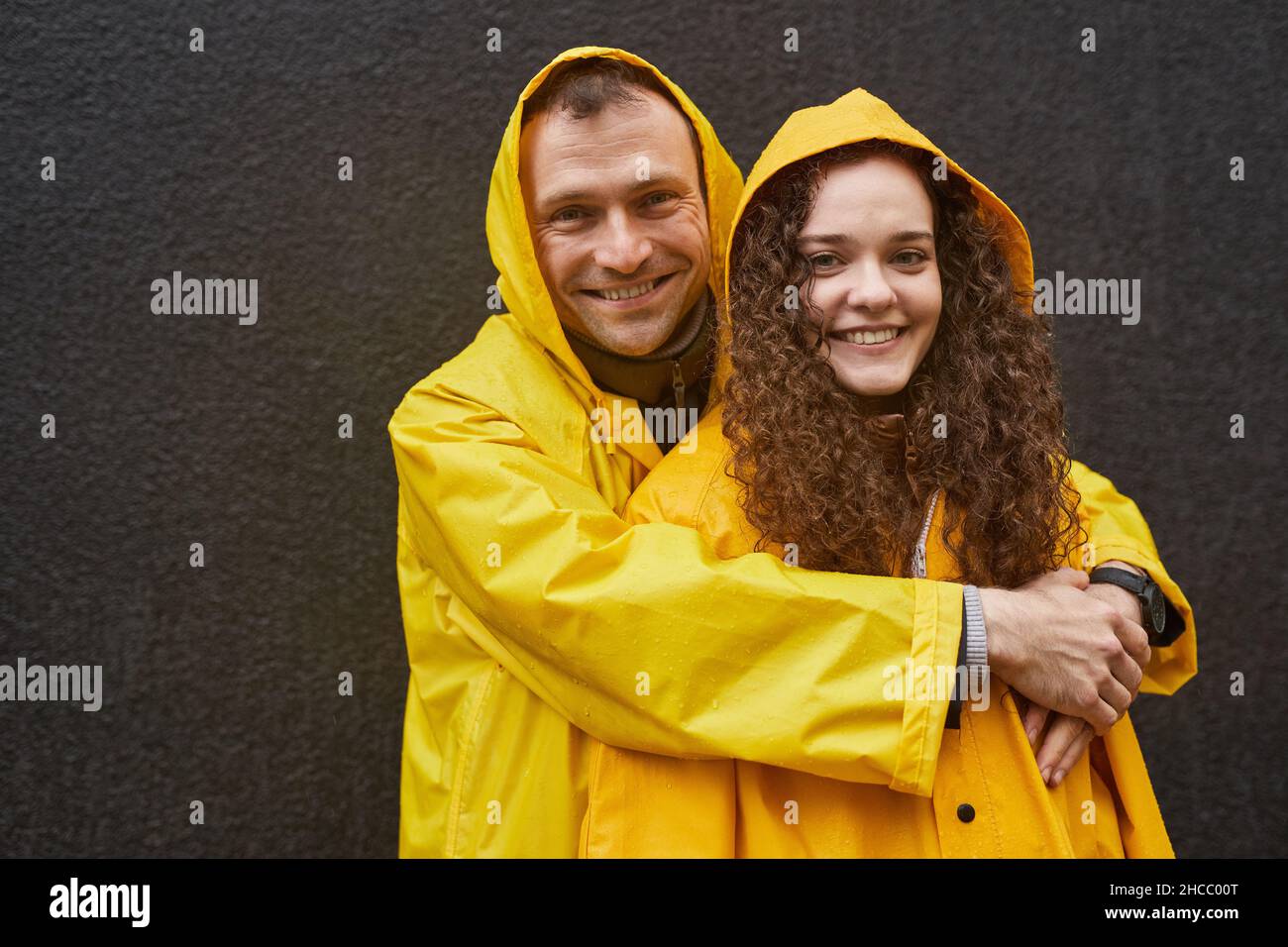 Portrait horizontal de l'homme et de la femme caucasiens amoureux portant des manteaux de pluie jaunes embrassant et regardant l'appareil photo Banque D'Images