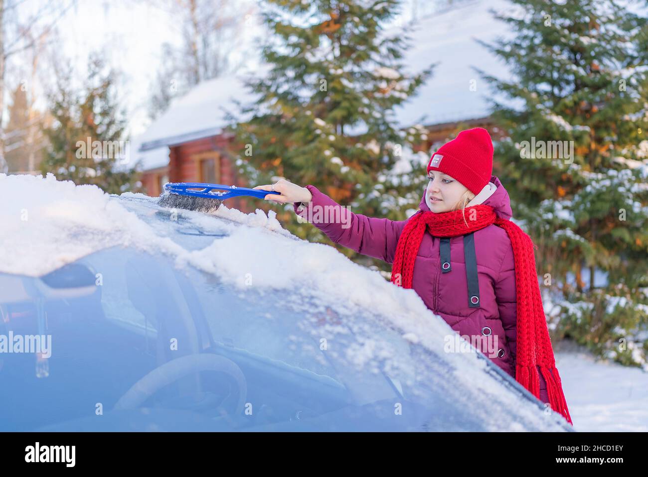 jeune femme dans un chapeau rouge nettoyant une voiture enneigée sur le fond d'une route d'hiver et le soleil couchant. Banque D'Images