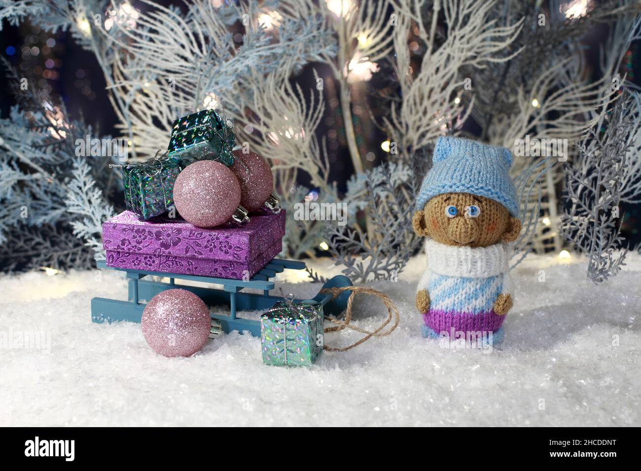 Un gnome tricoté en peluche se tient sur la neige artificielle et un traîneau en peluche avec des cadeaux se tient à côté de lui sur fond d'arbre artificiel blanc et bleu Banque D'Images