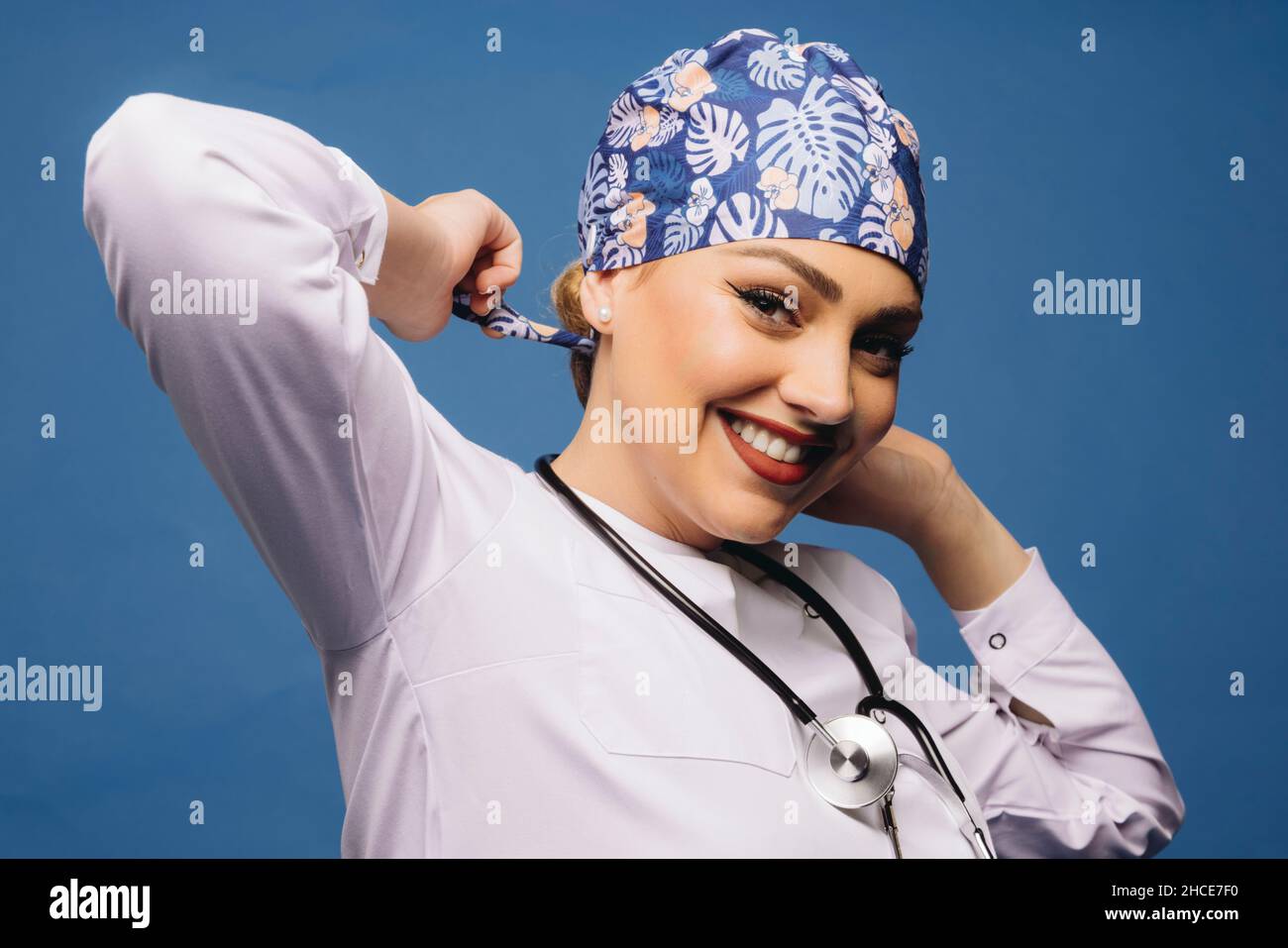 Gaie médecin femme portant une robe blanche médicale et stéthoscope nouant bandana uniforme sur la tête regardant la caméra Banque D'Images