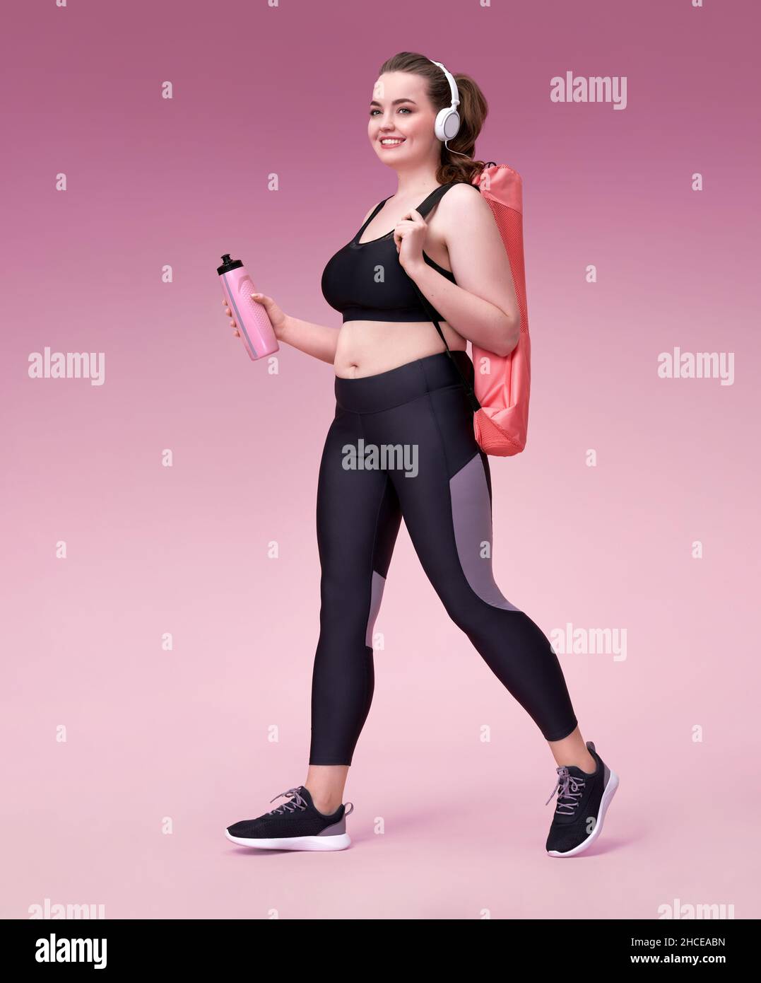 Allez à la formation !Femme sportive souriante avec tapis de yoga et bouteille d'eau.Photo du modèle avec une silhouette curvy dans des vêtements de sport tendance sur fond rose. Banque D'Images