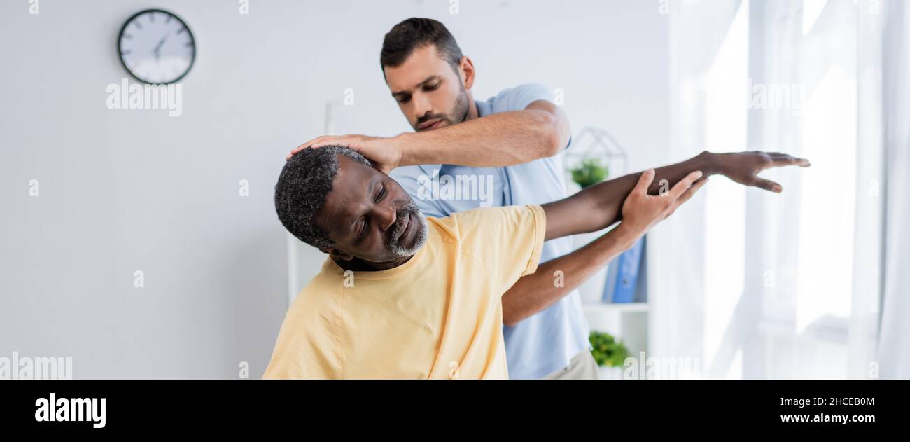 kinésithérapeute s'étirant bras de l'homme afro-américain tout en l'examinant dans le centre de réadaptation, bannière Banque D'Images