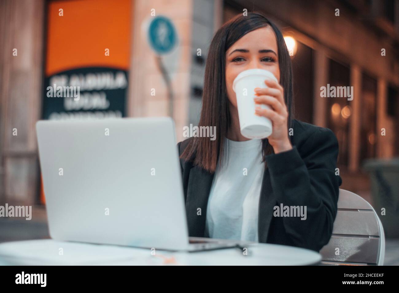Une jeune femme souriante qui boit un café dans un café de la ville tout en travaillant avec son ordinateur portable Banque D'Images