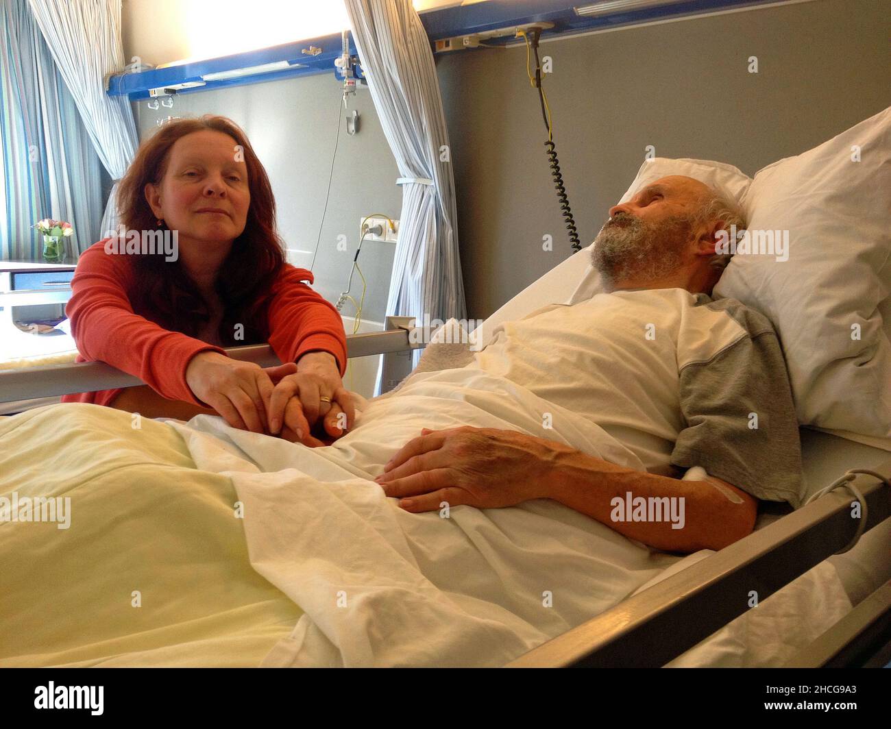 homme de 83 ans allongé dans un lit d'hôpital.Sa fille est assise à côté du lit et garde sa main. Banque D'Images