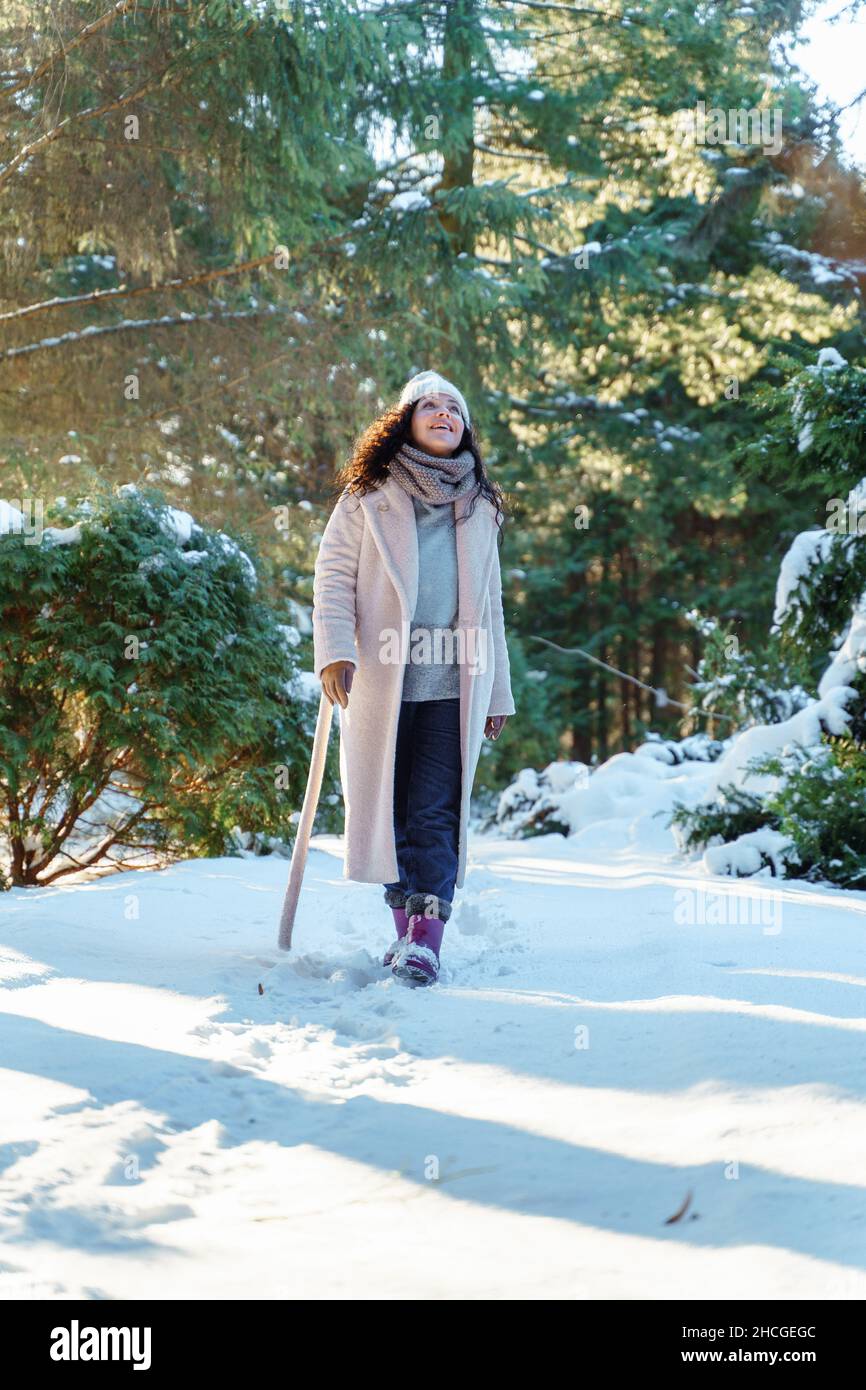 Souriante belle jeune femme avec de longues promenades à cheveux noirs regardant vers le haut portant des vêtements chauds devant le sapin dans la forêt enneigée d'hiver.Profitez de na Banque D'Images