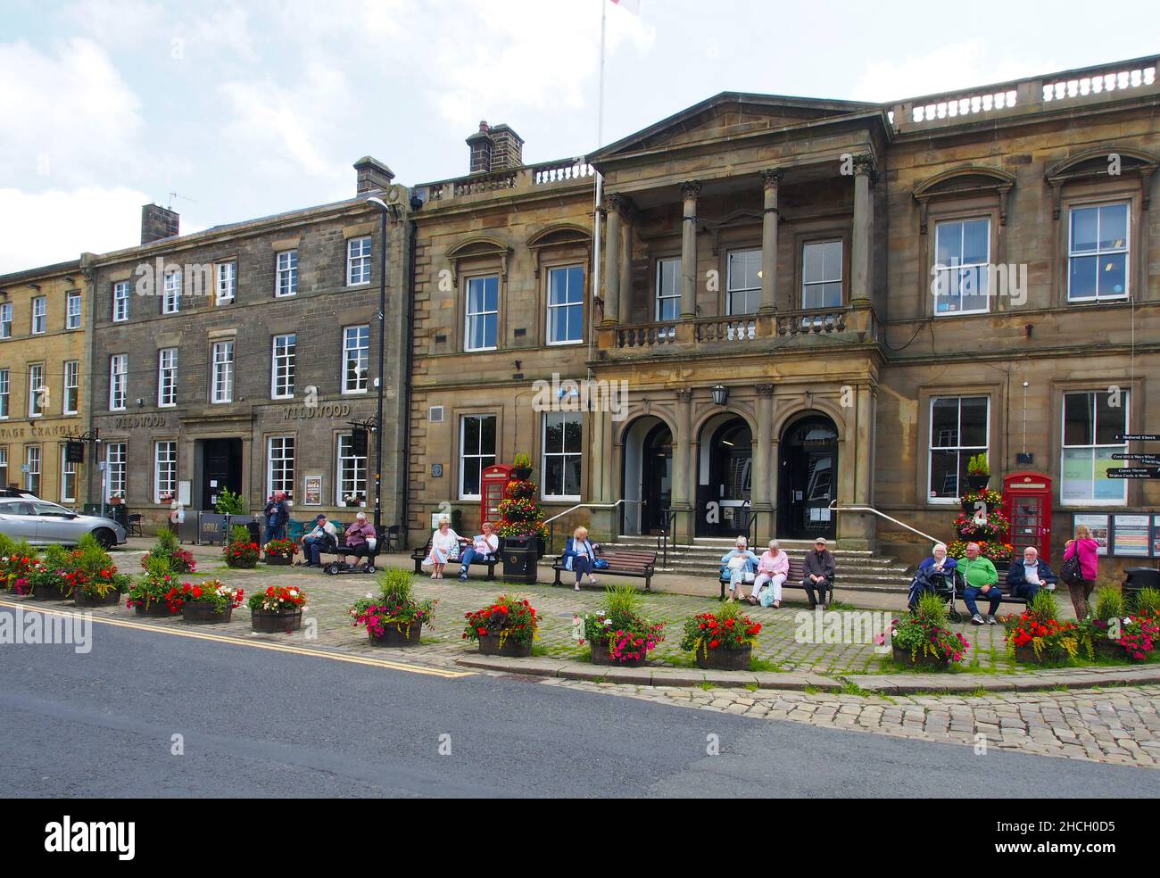 Les seniors profitent du soleil assis sur des bancs à l'extérieur de l'hôtel de ville de Skipton, dans le North Yorkshire, en Angleterre, avec des fleurs dans des baignoires au premier plan. Banque D'Images