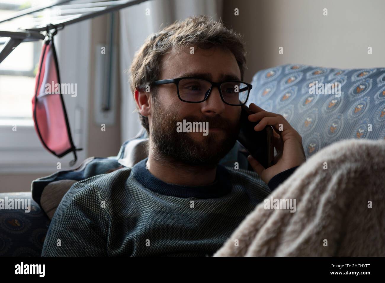 Un jeune homme, avec des verres de pâtes, barbu, parle sur le téléphone portable avec quelqu'un, tout en se reposant sur le canapé, couvert d'une couverture, dans l'apa partagée Banque D'Images