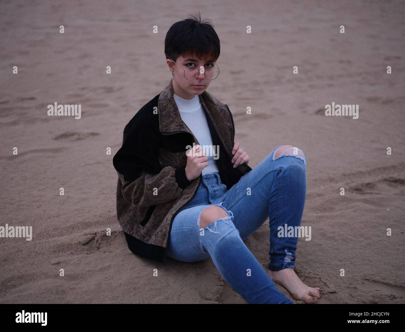 personne transgenre assise sur le sable avec une veste et des lunettes Banque D'Images