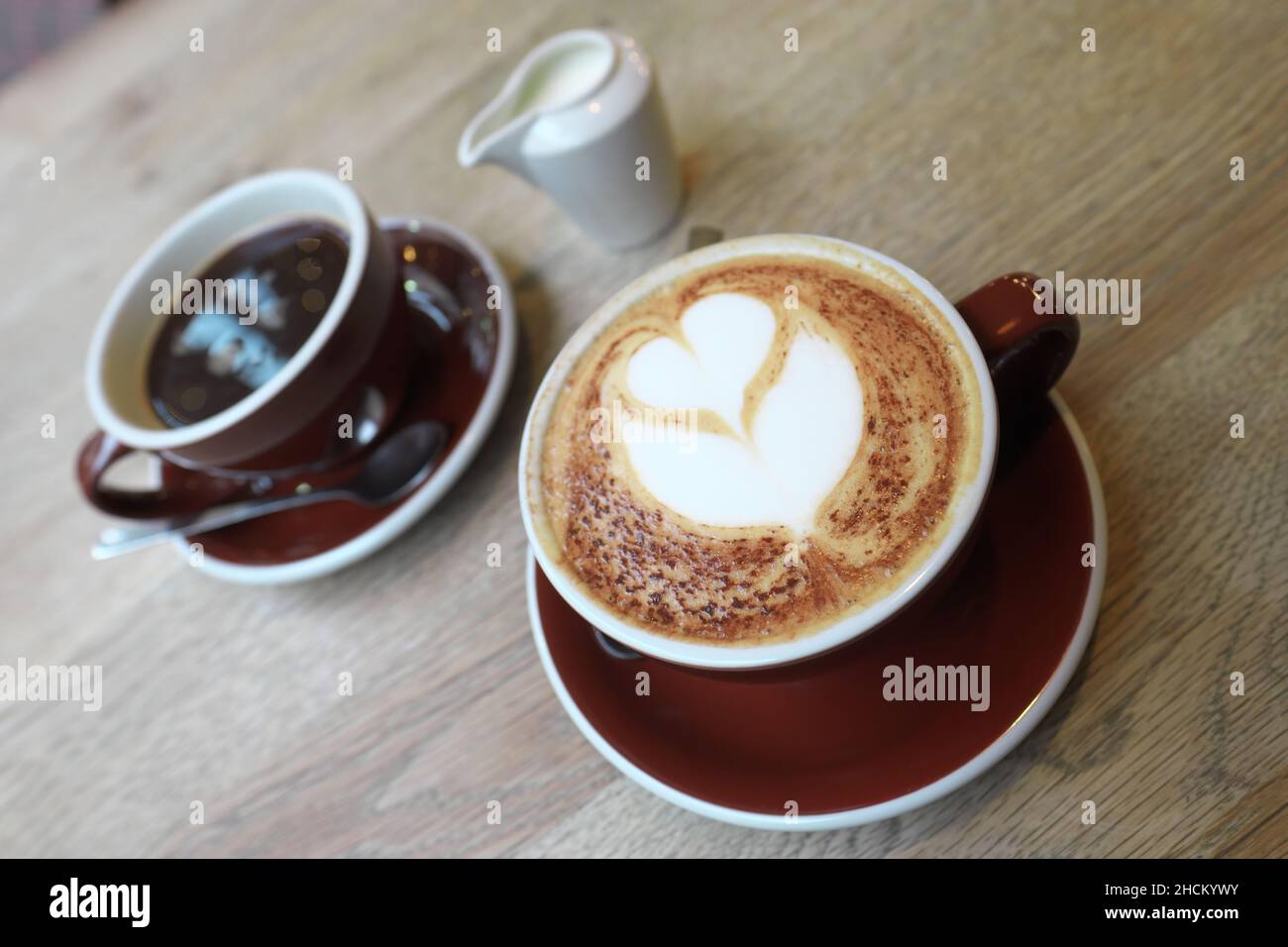 Un café Cappuccino avec un motif coeur décoré et un café noir avec un pot de lait derrière Banque D'Images