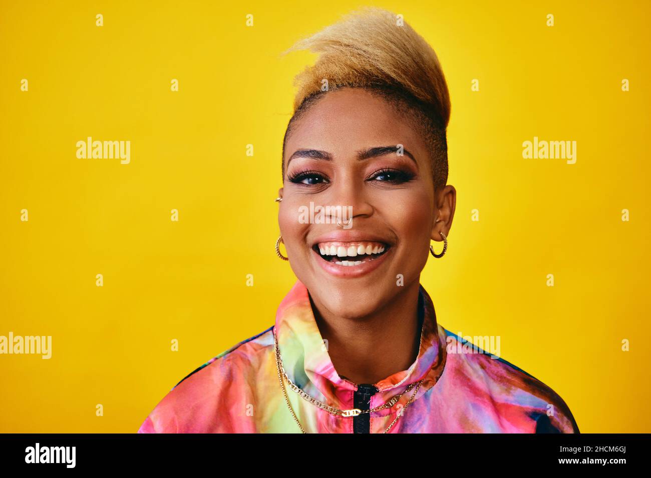 Bonne jeune femme élégante souriant avec des cheveux courts tendance, des bijoux et une veste colorée sur fond jaune Banque D'Images