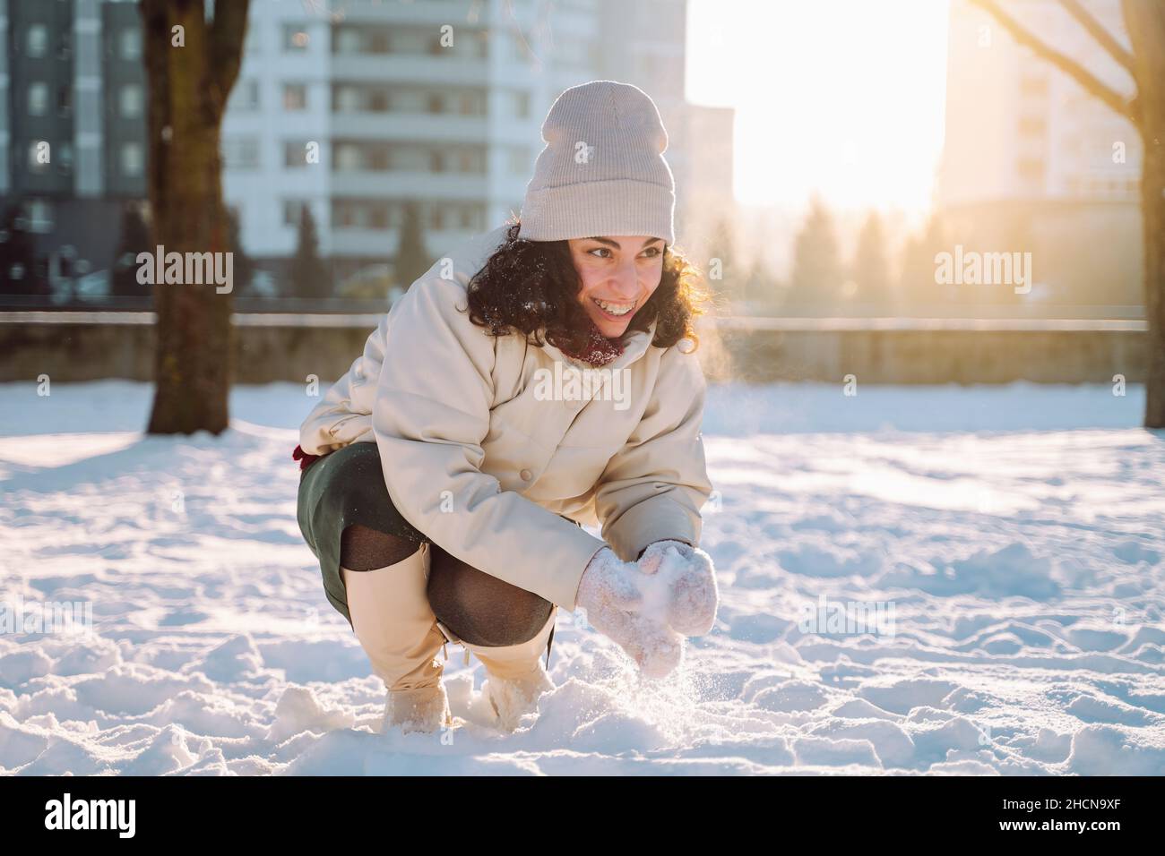 Souriante jeune femme se moque de jouer avec la neige en portant des vêtements blancs chauds dans un parc hivernal enneigé.Profiter de la nature, des vacances d'hiver, des émotions positives Banque D'Images
