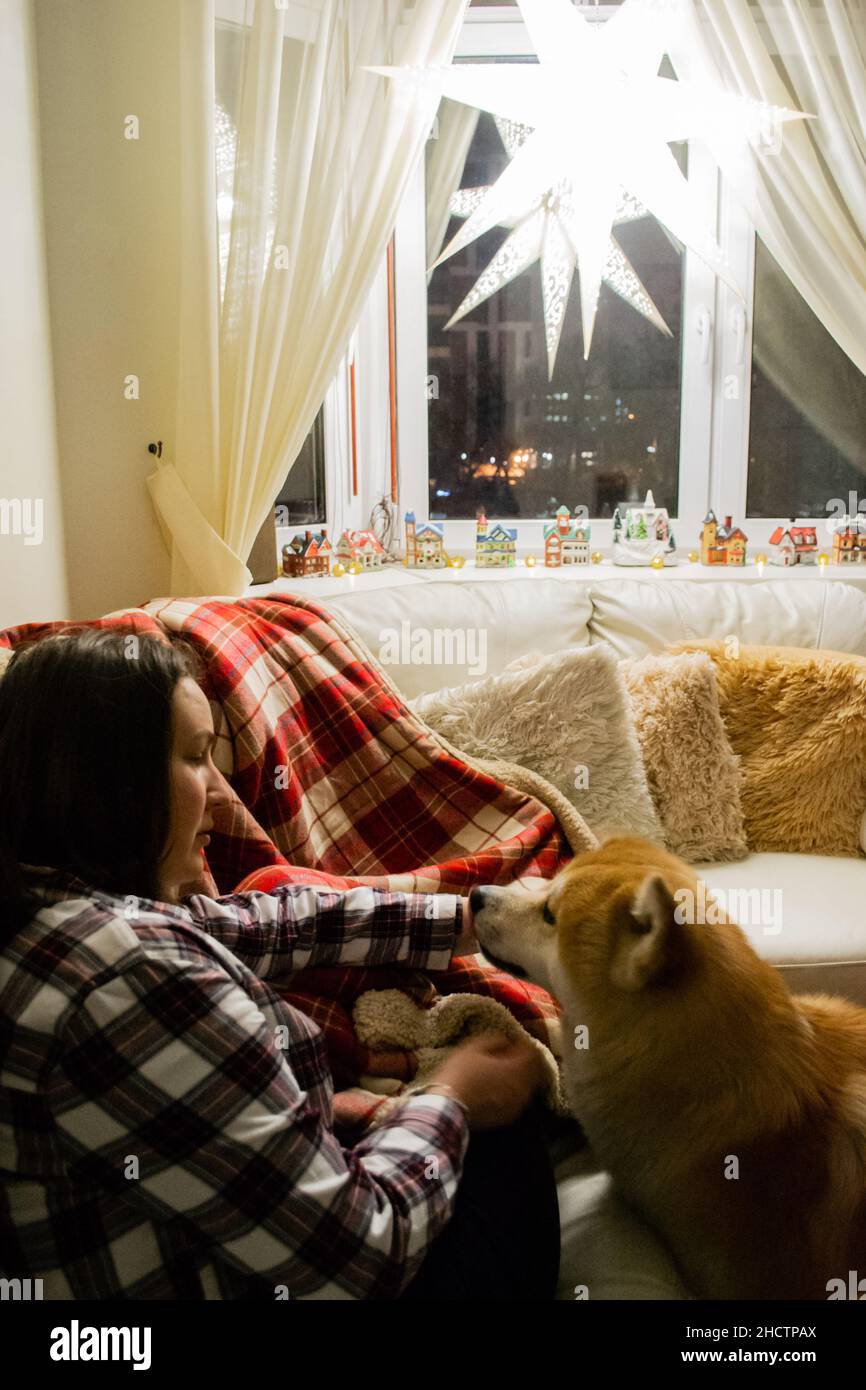 Une jeune femme sur un canapé avec un chien Akita près d'elle.Étoile de Noël sur la fenêtre: décoration de vacances.Maison confortable.Image verticale. Banque D'Images
