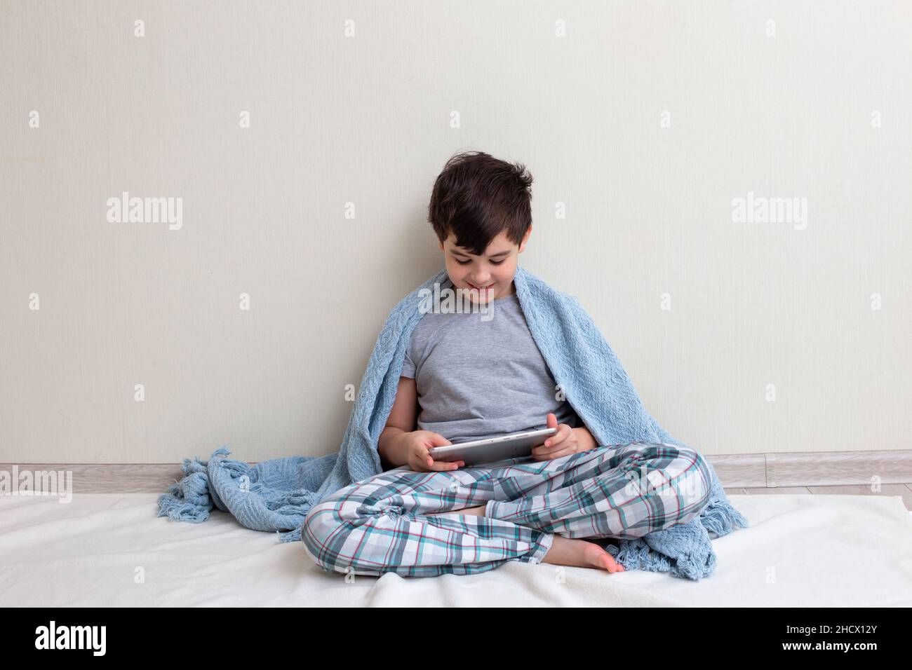 Un beau garçon, un adolescent en pyjama gris, sur le sol, sourit, utilise une tablette numérique Banque D'Images